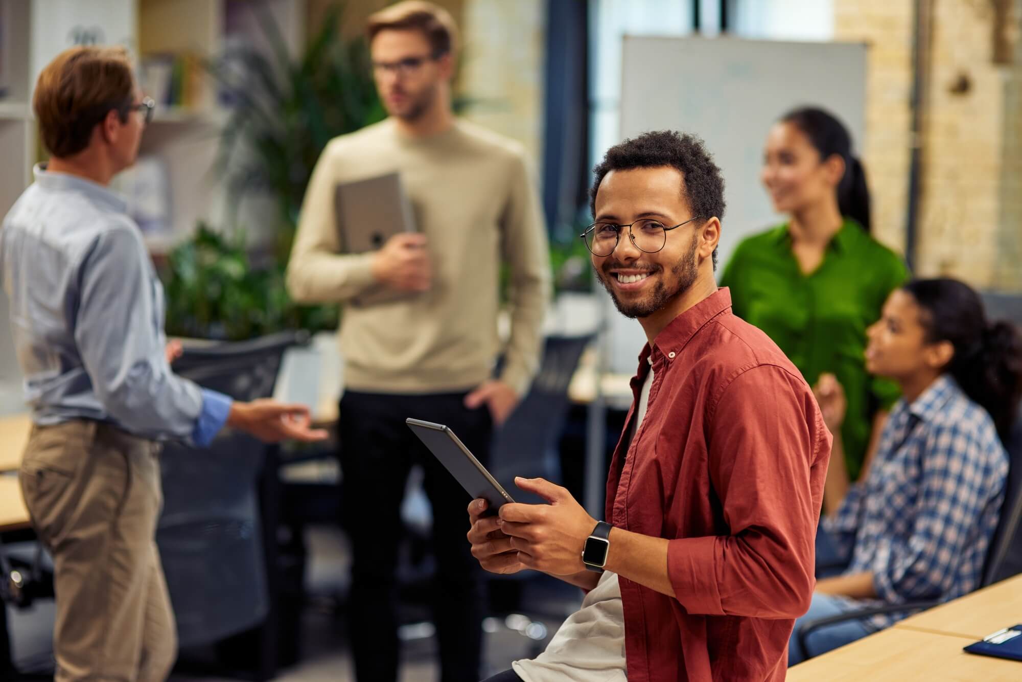 young-cheerful-mixed-race-man-holding-digital-tablet-and-smiling-at-camera-while-working-with.jpg